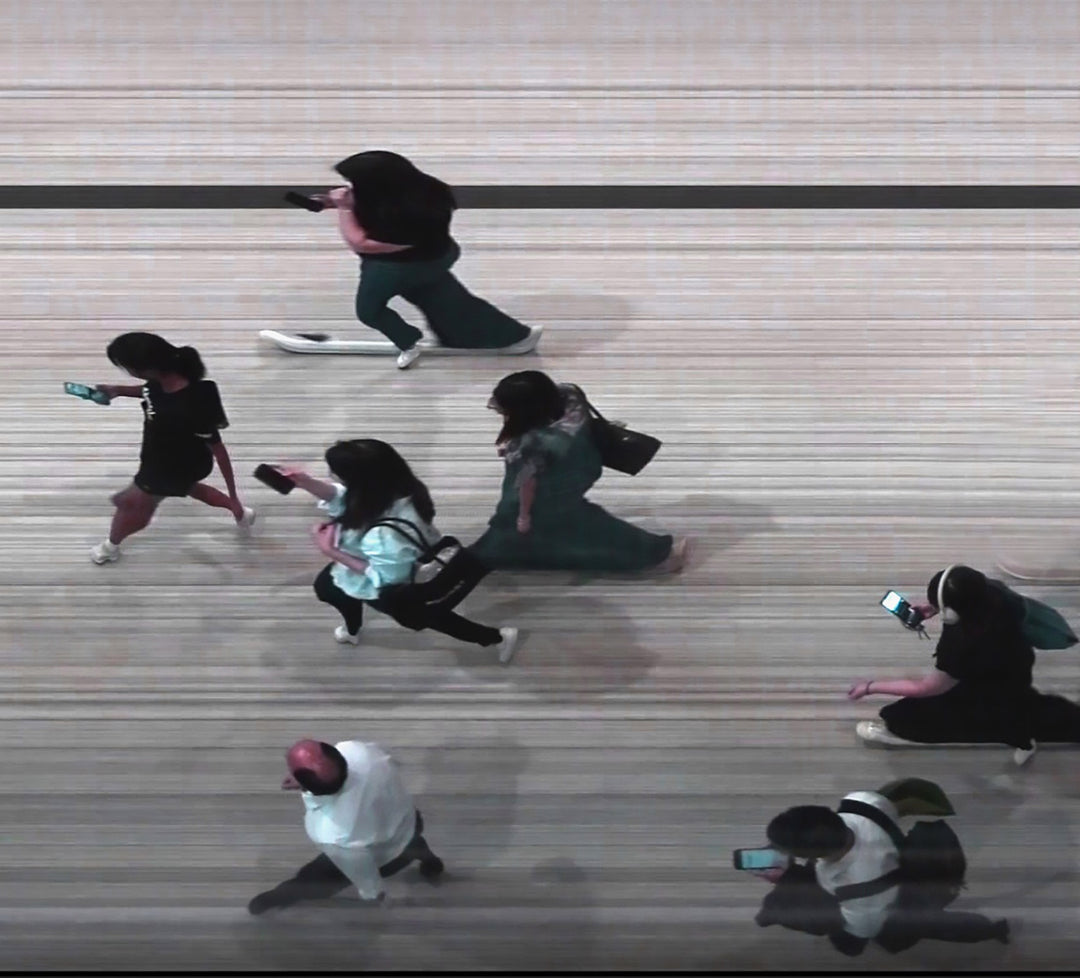 High-angle composite image of Hong Kong commuters moving through a brightly lit station, captured using high-speed video reconstruction. The figures are partially blurred and distorted, their faces downturned toward phones—creating a visual rhythm that blurs individuality, echoing the dehumanising pace of mass transit systems.