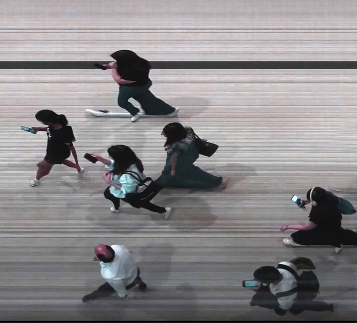 High-angle composite image of Hong Kong commuters moving through a brightly lit station, captured using high-speed video reconstruction. The figures are partially blurred and distorted, their faces downturned toward phones—creating a visual rhythm that blurs individuality, echoing the dehumanising pace of mass transit systems.
