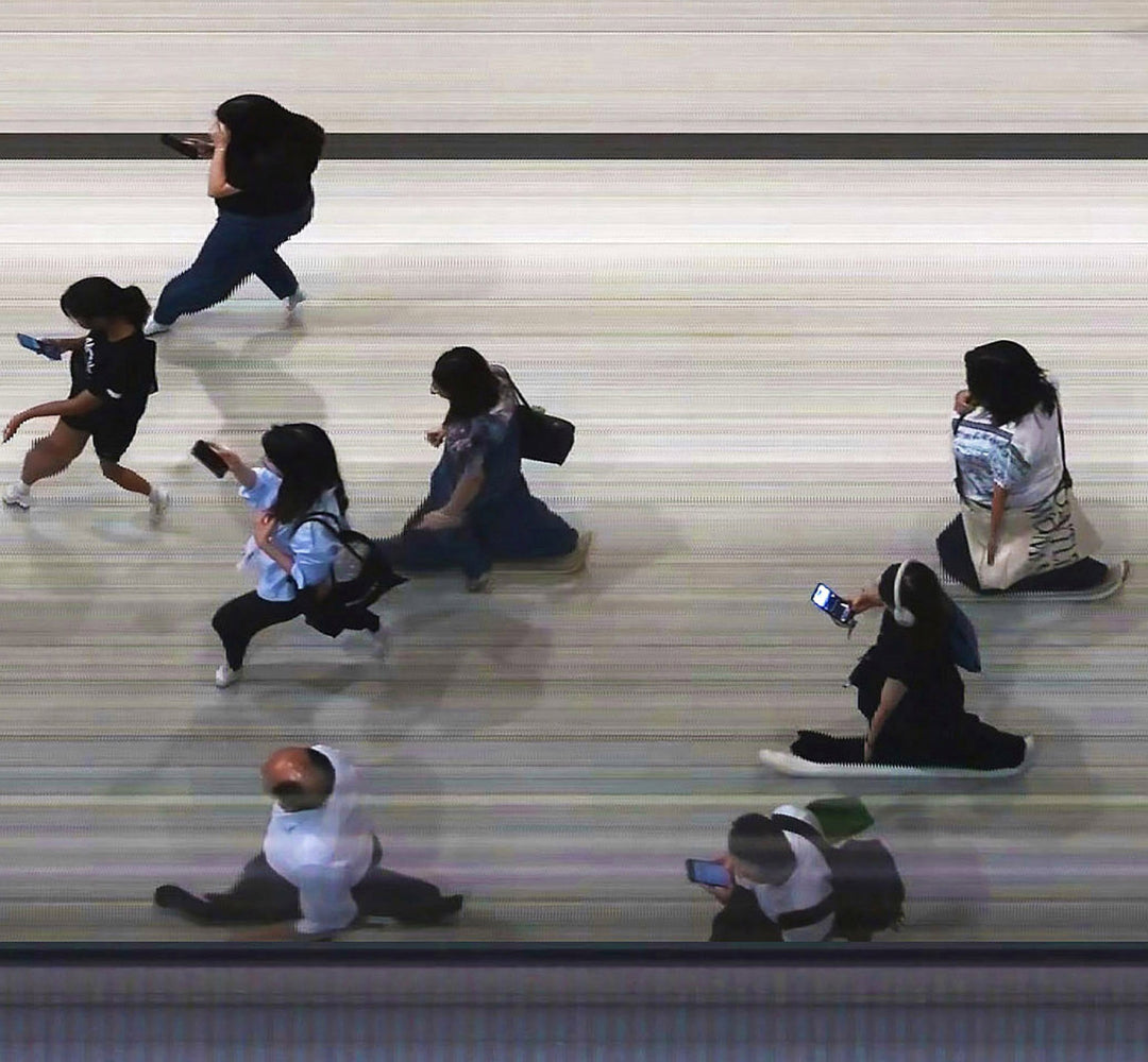 High-angle composite image of Hong Kong commuters moving through a brightly lit station, captured using high-speed video reconstruction. The figures are partially blurred and distorted, their faces downturned toward phones—creating a visual rhythm that blurs individuality, echoing the dehumanising pace of mass transit systems.