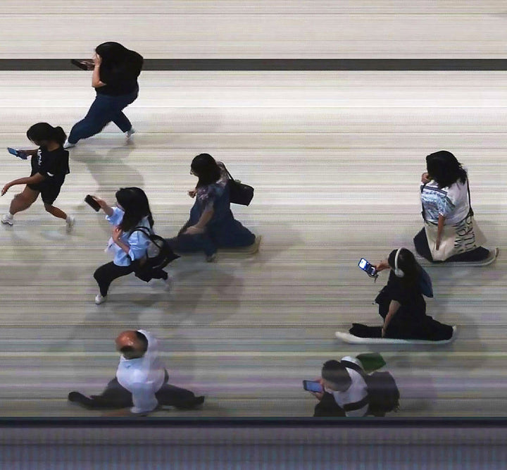 High-angle composite image of Hong Kong commuters moving through a brightly lit station, captured using high-speed video reconstruction. The figures are partially blurred and distorted, their faces downturned toward phones—creating a visual rhythm that blurs individuality, echoing the dehumanising pace of mass transit systems.