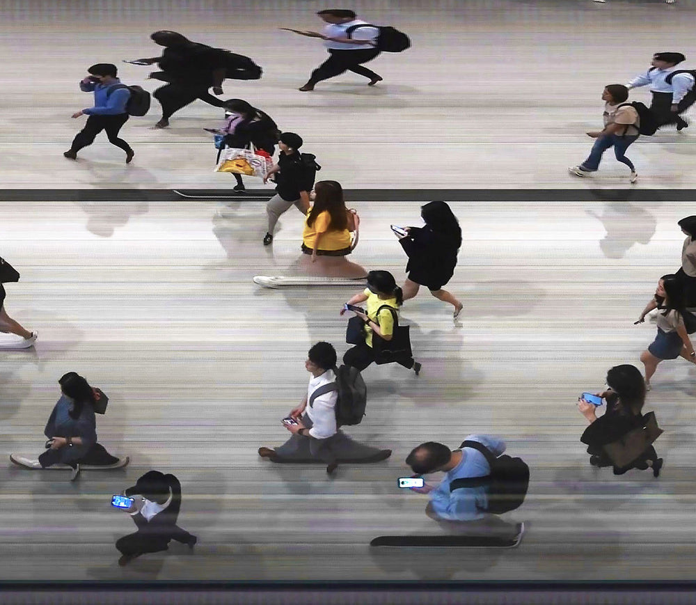 High-angle composite image of Hong Kong commuters moving through a brightly lit station, captured using high-speed video reconstruction. The figures are partially blurred and distorted, their faces downturned toward phones—creating a visual rhythm that blurs individuality, echoing the dehumanising pace of mass transit systems.