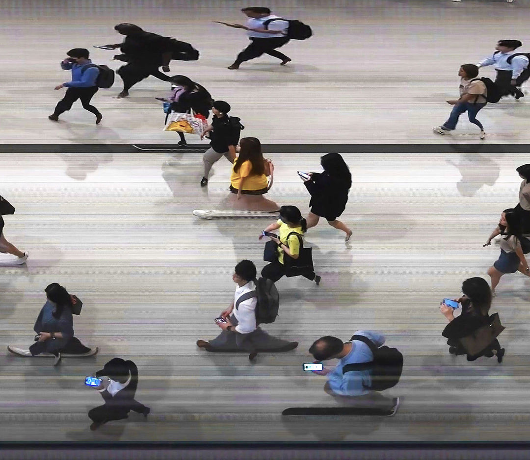 High-angle composite image of Hong Kong commuters moving through a brightly lit station, captured using high-speed video reconstruction. The figures are partially blurred and distorted, their faces downturned toward phones—creating a visual rhythm that blurs individuality, echoing the dehumanising pace of mass transit systems.
