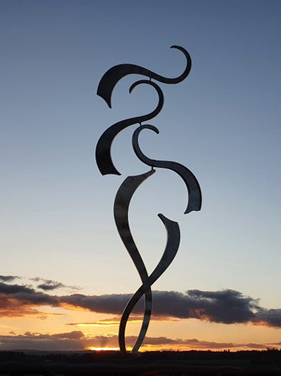 Graceful stainless steel sculpture with sweeping, curved forms twisting upward, set in a garden against a sunset
