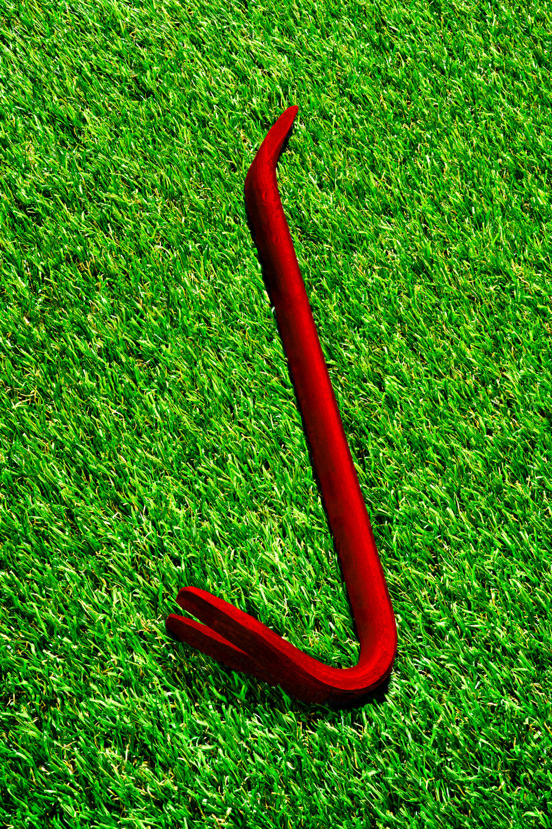 Photograph of a vivid red crowbar placed diagonally on a lush green grass background, contrasting industrial and organic textures.
