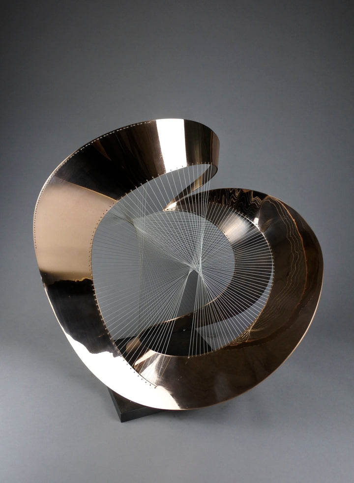 Curvilinear by Thomas Joynes – a unique commissioned sculpture in fabricated bronze sheet with mirror polished and patinated finishes. The work features a dynamic, flowing bronze form intersected by a finely woven matrix of tensioned wire. Balanced on a black stone plinth, this complex yet graceful piece was created for the One Blackfriars luxury development in London, echoing the architectural fluidity of the building with a contemporary nod to Hepworth. Photographed against a soft grey backdrop.