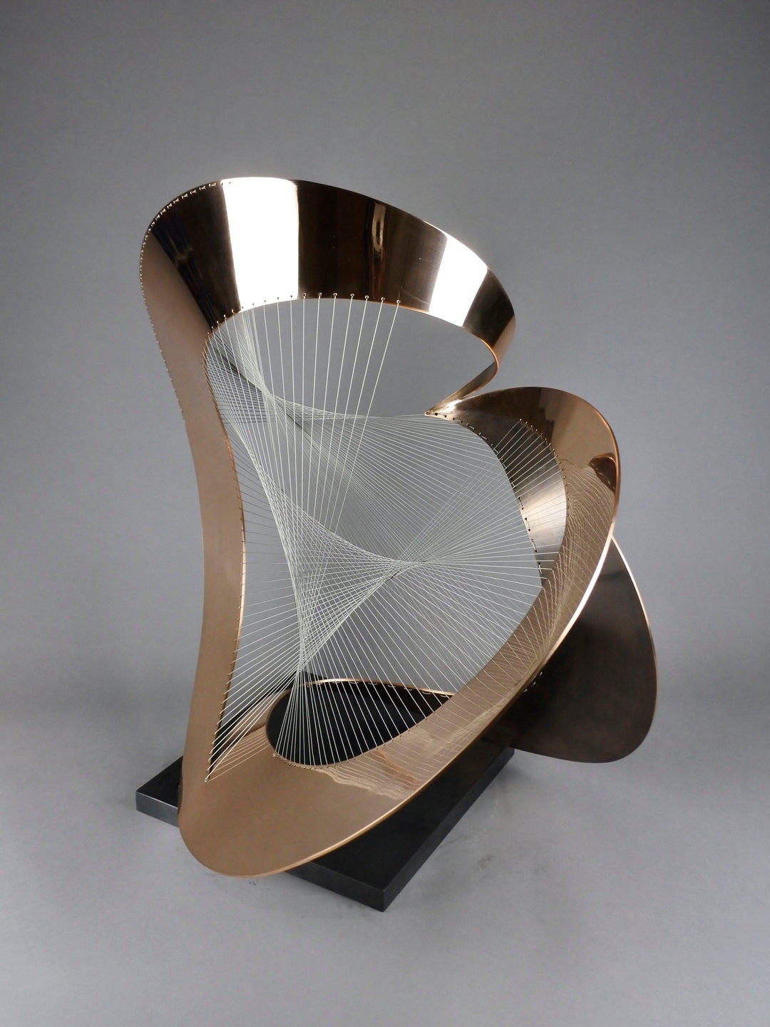 Curvilinear by Thomas Joynes – a unique commissioned sculpture in fabricated bronze sheet with mirror polished and patinated finishes. The work features a dynamic, flowing bronze form intersected by a finely woven matrix of tensioned wire. Balanced on a black stone plinth, this complex yet graceful piece was created for the One Blackfriars luxury development in London, echoing the architectural fluidity of the building with a contemporary nod to Hepworth. Photographed against a soft grey backdrop.