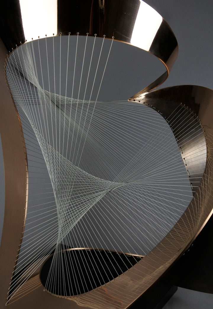Curvilinear by Thomas Joynes – a unique commissioned sculpture in fabricated bronze sheet with mirror polished and patinated finishes. The work features a dynamic, flowing bronze form intersected by a finely woven matrix of tensioned wire. Balanced on a black stone plinth, this complex yet graceful piece was created for the One Blackfriars luxury development in London, echoing the architectural fluidity of the building with a contemporary nod to Hepworth. Photographed against a soft grey backdrop.