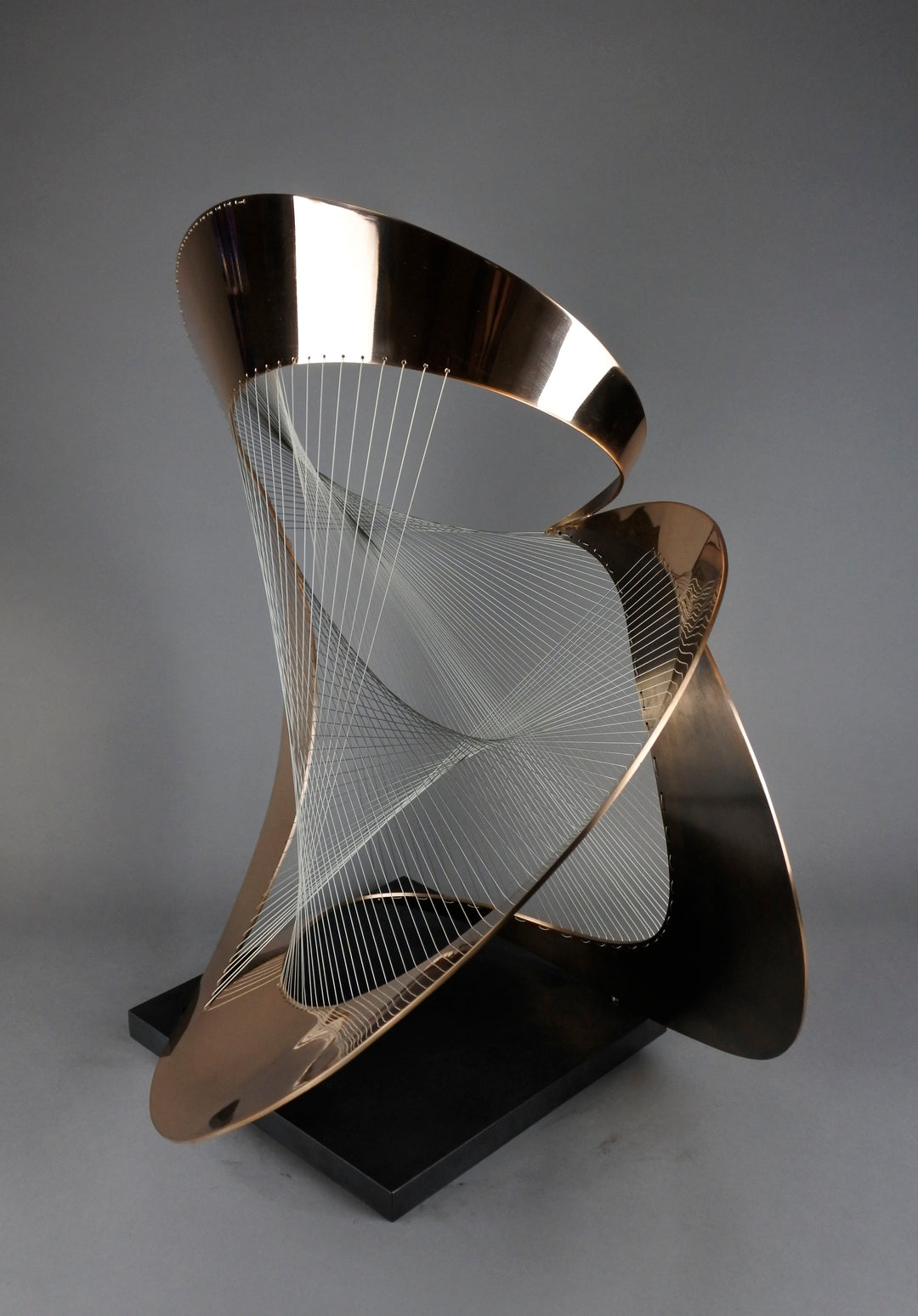 Curvilinear by Thomas Joynes – a unique commissioned sculpture in fabricated bronze sheet with mirror polished and patinated finishes. The work features a dynamic, flowing bronze form intersected by a finely woven matrix of tensioned wire. Balanced on a black stone plinth, this complex yet graceful piece was created for the One Blackfriars luxury development in London, echoing the architectural fluidity of the building with a contemporary nod to Hepworth. Photographed against a soft grey backdrop.
