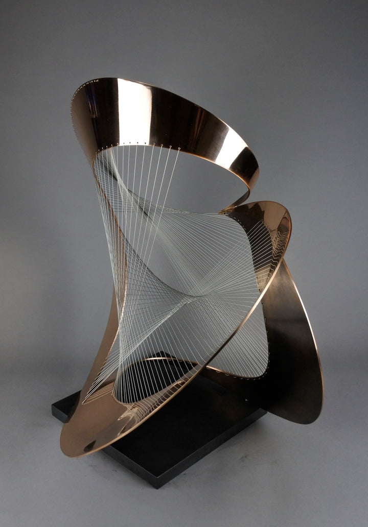 Curvilinear by Thomas Joynes – a unique commissioned sculpture in fabricated bronze sheet with mirror polished and patinated finishes. The work features a dynamic, flowing bronze form intersected by a finely woven matrix of tensioned wire. Balanced on a black stone plinth, this complex yet graceful piece was created for the One Blackfriars luxury development in London, echoing the architectural fluidity of the building with a contemporary nod to Hepworth. Photographed against a soft grey backdrop.