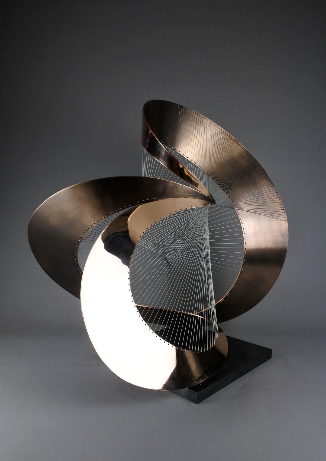 Curvilinear by Thomas Joynes – a unique commissioned sculpture in fabricated bronze sheet with mirror polished and patinated finishes. The work features a dynamic, flowing bronze form intersected by a finely woven matrix of tensioned wire. Balanced on a black stone plinth, this complex yet graceful piece was created for the One Blackfriars luxury development in London, echoing the architectural fluidity of the building with a contemporary nod to Hepworth. Photographed against a soft grey backdrop.