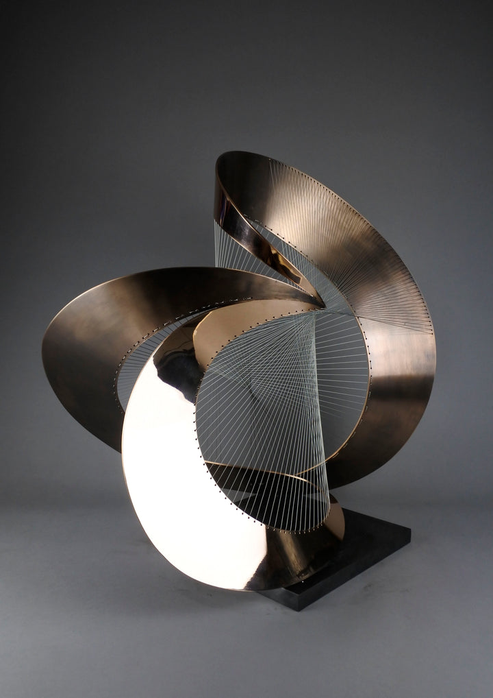 Curvilinear by Thomas Joynes – a unique commissioned sculpture in fabricated bronze sheet with mirror polished and patinated finishes. The work features a dynamic, flowing bronze form intersected by a finely woven matrix of tensioned wire. Balanced on a black stone plinth, this complex yet graceful piece was created for the One Blackfriars luxury development in London, echoing the architectural fluidity of the building with a contemporary nod to Hepworth. Photographed against a soft grey backdrop.