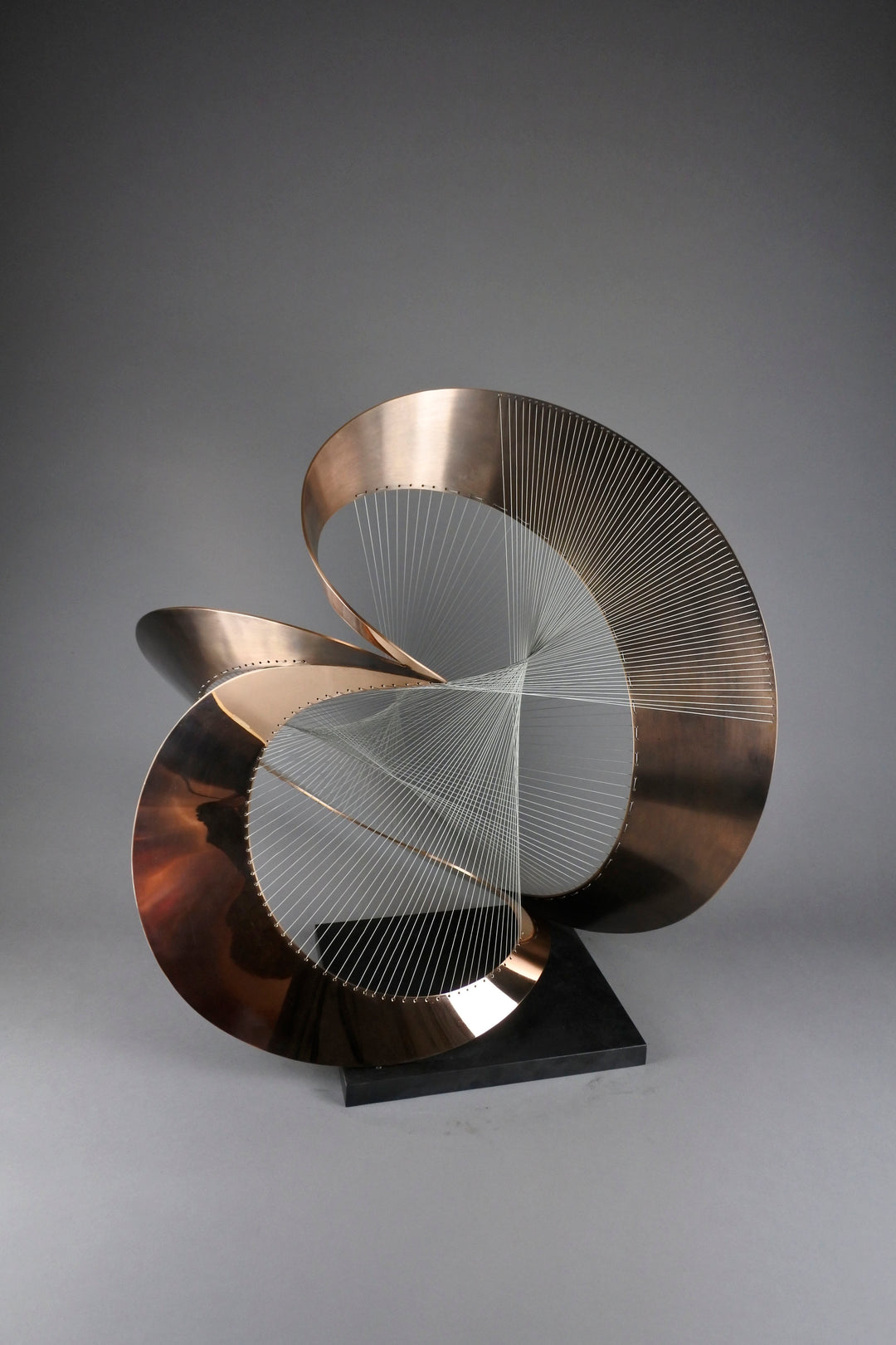 Curvilinear by Thomas Joynes – a unique commissioned sculpture in fabricated bronze sheet with mirror polished and patinated finishes. The work features a dynamic, flowing bronze form intersected by a finely woven matrix of tensioned wire. Balanced on a black stone plinth, this complex yet graceful piece was created for the One Blackfriars luxury development in London, echoing the architectural fluidity of the building with a contemporary nod to Hepworth. Photographed against a soft grey backdrop.