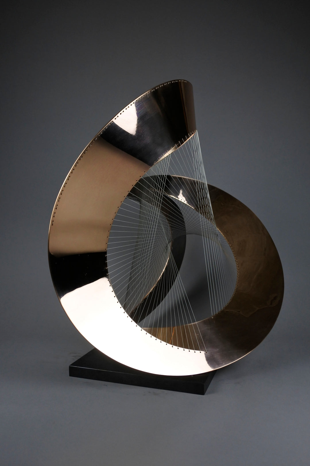 Curvilinear by Thomas Joynes – a unique commissioned sculpture in fabricated bronze sheet with mirror polished and patinated finishes. The work features a dynamic, flowing bronze form intersected by a finely woven matrix of tensioned wire. Balanced on a black stone plinth, this complex yet graceful piece was created for the One Blackfriars luxury development in London, echoing the architectural fluidity of the building with a contemporary nod to Hepworth. Photographed against a soft grey backdrop.