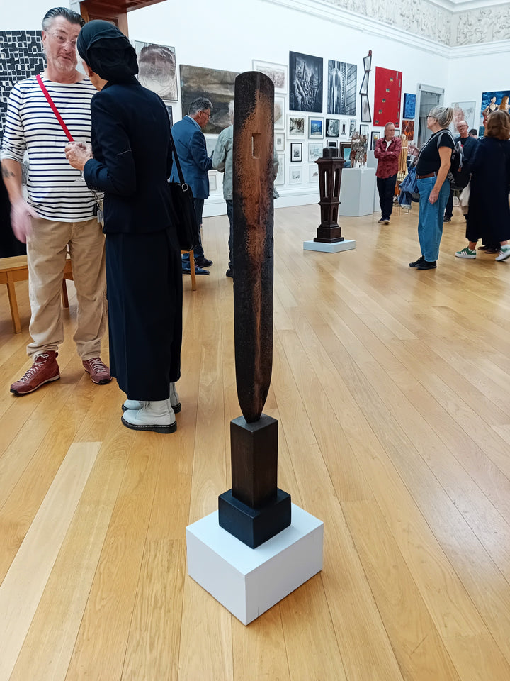 Tall, slender carved elm sculpture standing on an oak base, shaped like an abstracted Stone Age axe with a dark, textured lower section and a narrow architectural opening near the top, creating a quiet, contemplative presence that balances weight, history, and calm in a minimal indoor setting.