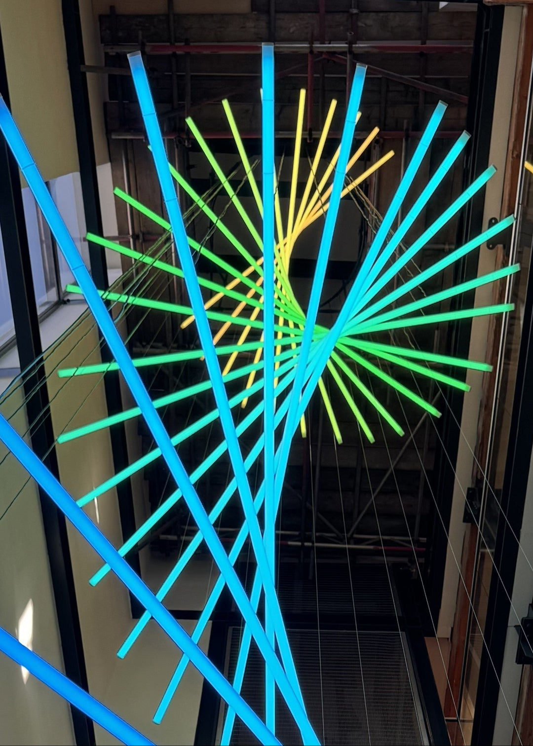 Suspended spiral light installation composed of intersecting illuminated linear elements arranged around a vertical stairwell void, with coloured light bars radiating and twisting through space to echo the geometry and movement of the surrounding architecture, creating layered reflections and depth within the entrance atrium.