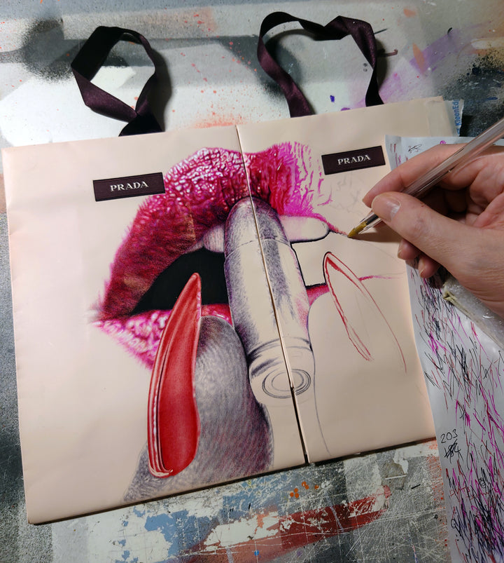 Original artwork titled “Apply With Caution” by James Mylne, featuring a provocative hyperrealistic drawing of glossy red lips applying lipstick formed from a bullet. Created using red and black ballpoint pens and acrylic marker on two Prada gift bags, the work is framed in a black box frame with red detailing and ribbon handles exposed.