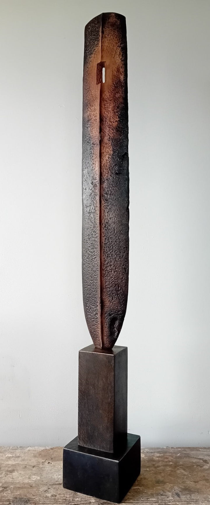 Tall, slender carved elm sculpture standing on an oak base, shaped like an abstracted Stone Age axe with a dark, textured lower section and a narrow architectural opening near the top, creating a quiet, contemplative presence that balances weight, history, and calm in a minimal indoor setting.