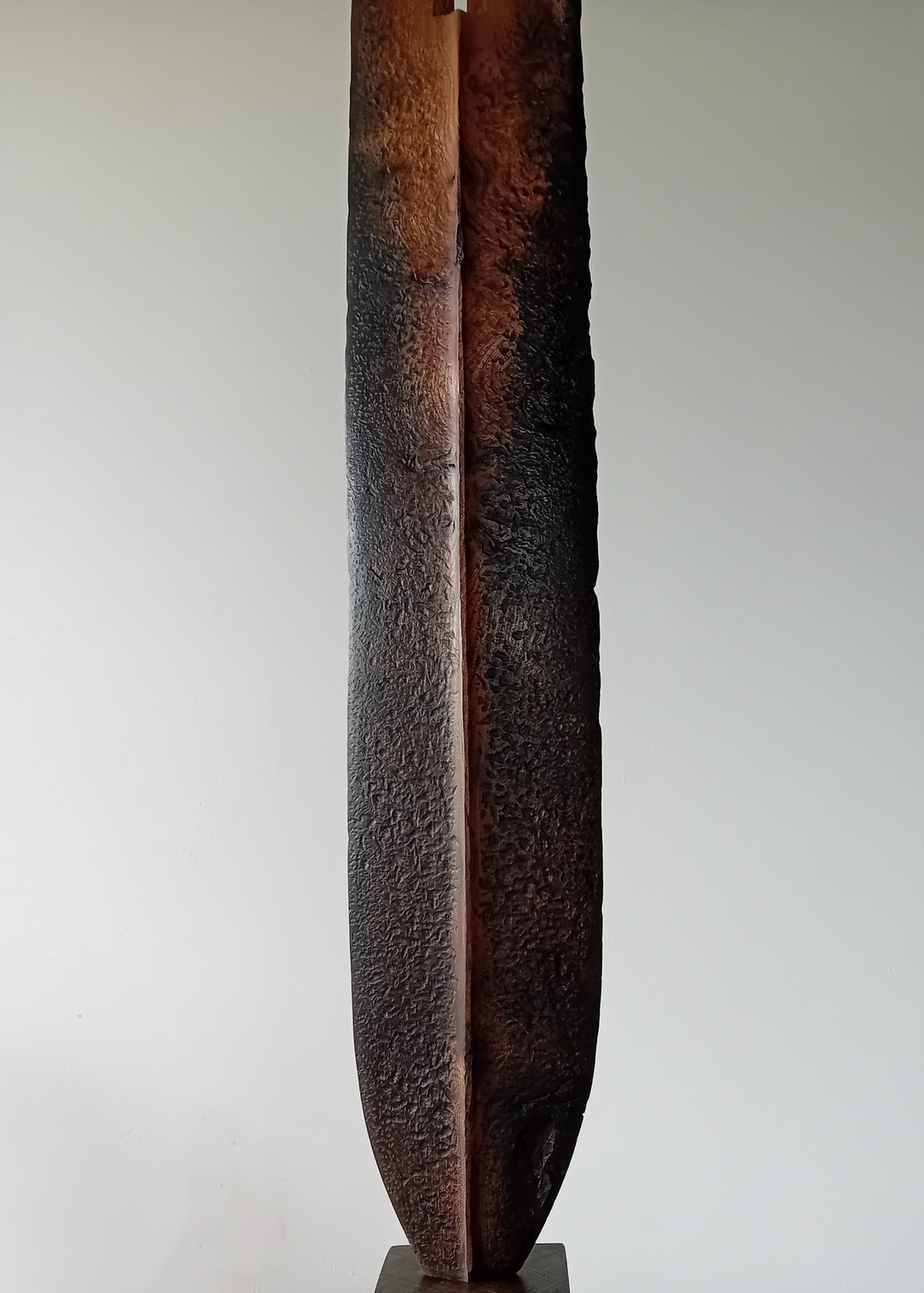 Tall, slender carved elm sculpture standing on an oak base, shaped like an abstracted Stone Age axe with a dark, textured lower section and a narrow architectural opening near the top, creating a quiet, contemplative presence that balances weight, history, and calm in a minimal indoor setting.