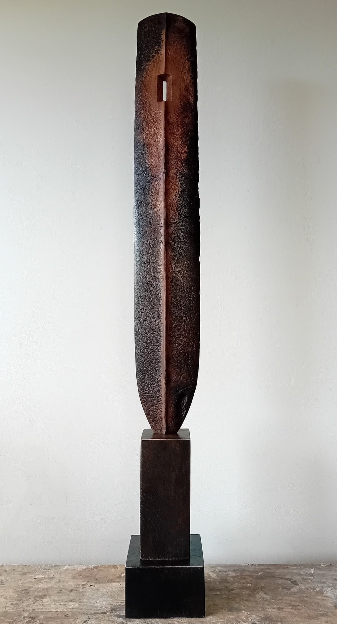 Tall, slender carved elm sculpture standing on an oak base, shaped like an abstracted Stone Age axe with a dark, textured lower section and a narrow architectural opening near the top, creating a quiet, contemplative presence that balances weight, history, and calm in a minimal indoor setting.
