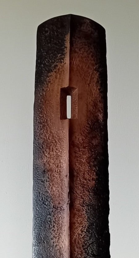 Tall, slender carved elm sculpture standing on an oak base, shaped like an abstracted Stone Age axe with a dark, textured lower section and a narrow architectural opening near the top, creating a quiet, contemplative presence that balances weight, history, and calm in a minimal indoor setting.