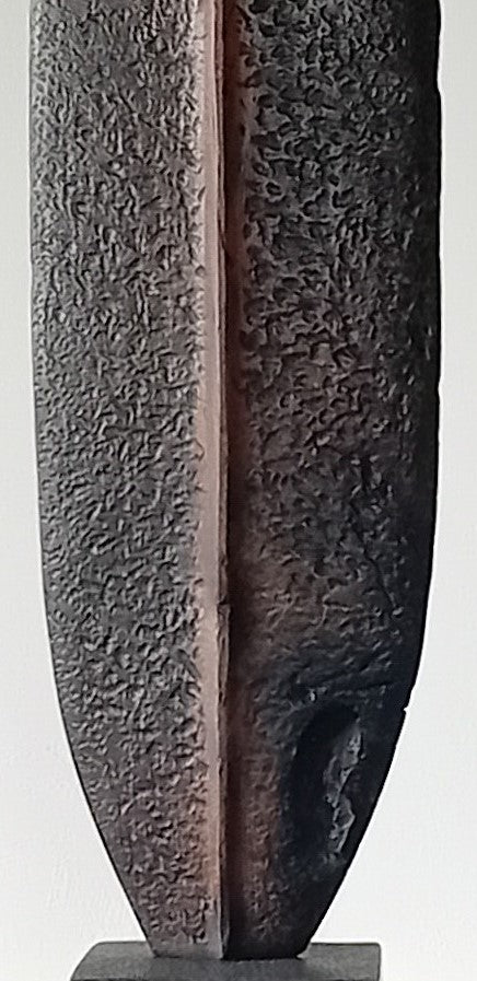 Tall, slender carved elm sculpture standing on an oak base, shaped like an abstracted Stone Age axe with a dark, textured lower section and a narrow architectural opening near the top, creating a quiet, contemplative presence that balances weight, history, and calm in a minimal indoor setting.