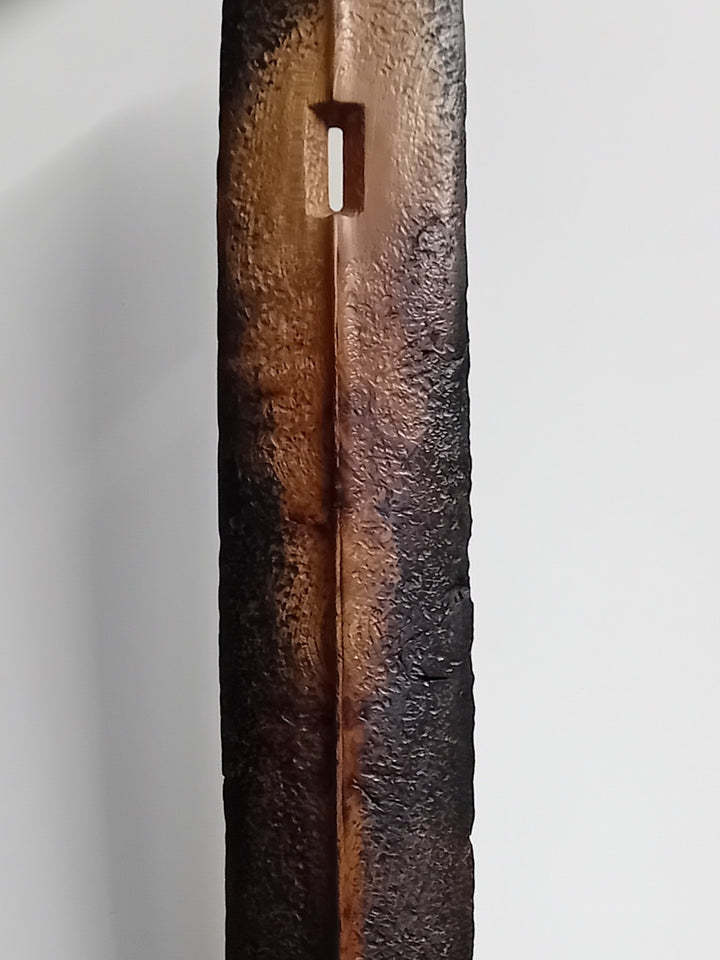 Tall, slender carved elm sculpture standing on an oak base, shaped like an abstracted Stone Age axe with a dark, textured lower section and a narrow architectural opening near the top, creating a quiet, contemplative presence that balances weight, history, and calm in a minimal indoor setting.