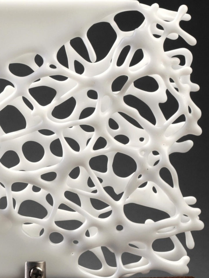 Diaphaneity VIII by Tracy Nicholls — a one-off kiln-formed glass and marble sculpture. Two panels of opaque white glass rest on a marble base, their surface pierced by an intricate lace-like web that seems to dissolve into open space. The organic strands ripple with gentle movement, allowing light to filter through and cast shifting shadows across surrounding surfaces, transforming solidity into a play of fragility and air.