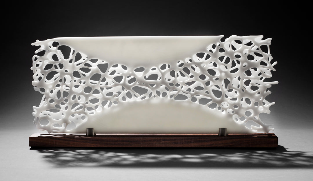 Diaphaneity VIII by Tracy Nicholls — a one-off kiln-formed glass and marble sculpture. Two panels of opaque white glass rest on a marble base, their surface pierced by an intricate lace-like web that seems to dissolve into open space. The organic strands ripple with gentle movement, allowing light to filter through and cast shifting shadows across surrounding surfaces, transforming solidity into a play of fragility and air.