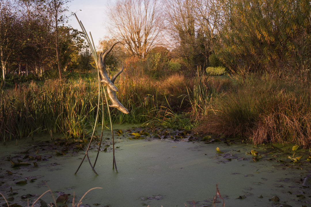 Simon Gudgeon bronze sculpture Diving Otters featuring two elegant diving otter forms suspended amongst tall reed-like structures within a landscaped waterside garden setting.