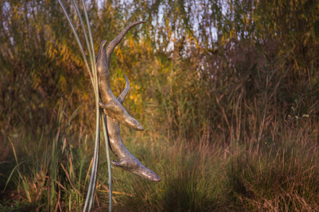 Simon Gudgeon bronze sculpture Diving Otters featuring two elegant diving otter forms suspended amongst tall reed-like structures within a landscaped waterside garden setting.