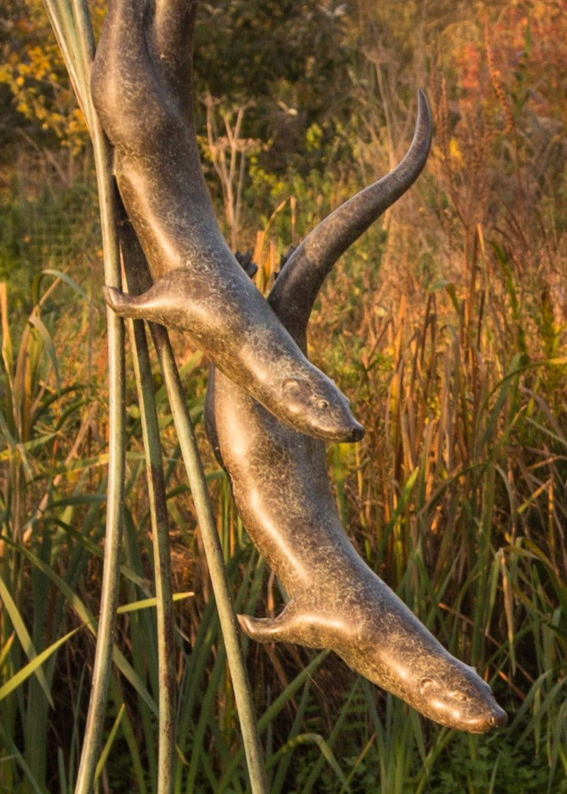 Simon Gudgeon bronze sculpture Diving Otters featuring two elegant diving otter forms suspended amongst tall reed-like structures within a landscaped waterside garden setting.
