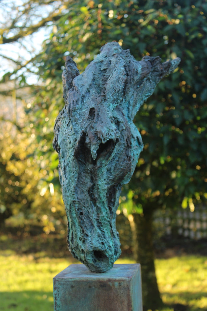 Abstract bronze sculpture of a horse’s head, mounted on a tall corten steel plinth, displayed in various garden and outdoor settings. The textured verdigris patina highlights the expressive contours and emotional form of the equine subject.