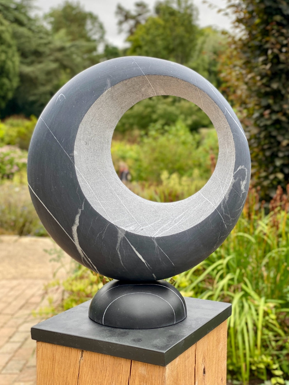 Abstract stone sculpture Eclipse – 2025 by Hamish Holman, carved from Nero Blue marble with a smooth circular form and hollowed centre, featuring natural white veining and a polished dark blue-grey surface, mounted on a wooden plinth.