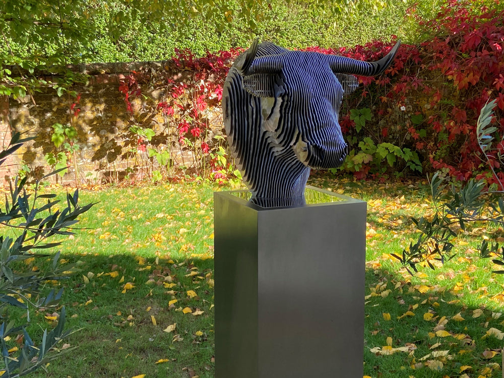 A bold stainless steel sculpture of a bull’s head by Tom Hiscox, composed of layered contour slices with curved horns and muscular form, set against vivid autumn foliage in an outdoor garden setting.
