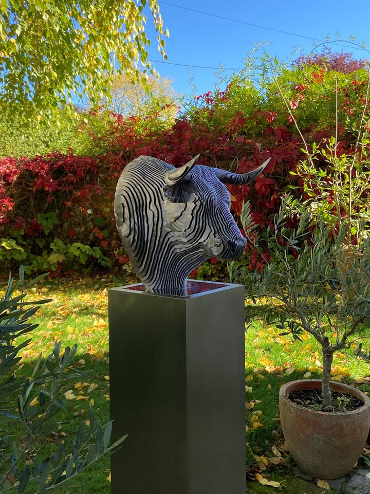 A bold stainless steel sculpture of a bull’s head by Tom Hiscox, composed of layered contour slices with curved horns and muscular form, set against vivid autumn foliage in an outdoor garden setting.