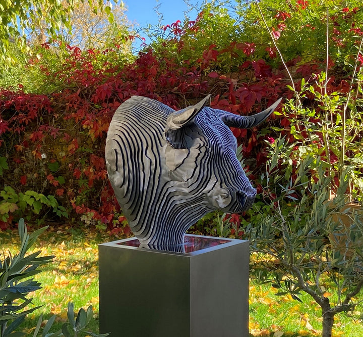 A bold stainless steel sculpture of a bull’s head by Tom Hiscox, composed of layered contour slices with curved horns and muscular form, set against vivid autumn foliage in an outdoor garden setting.