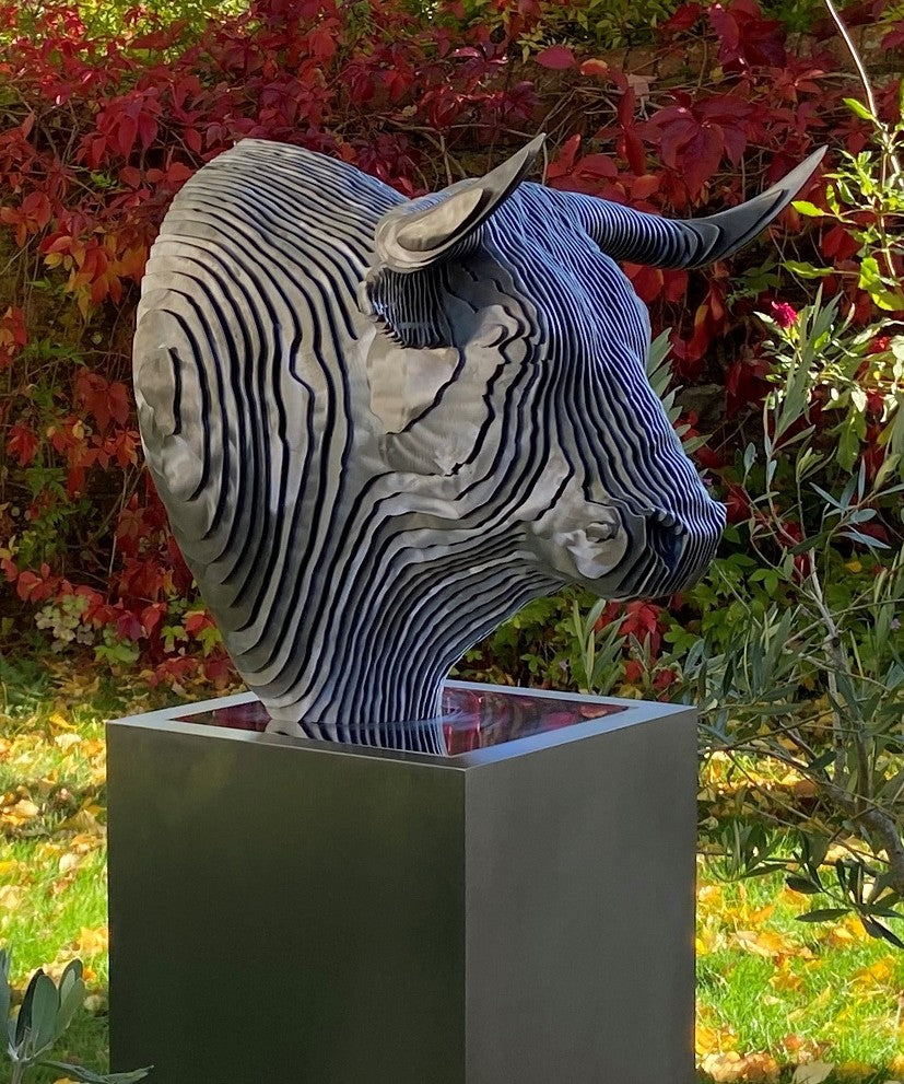 A bold stainless steel sculpture of a bull’s head by Tom Hiscox, composed of layered contour slices with curved horns and muscular form, set against vivid autumn foliage in an outdoor garden setting.