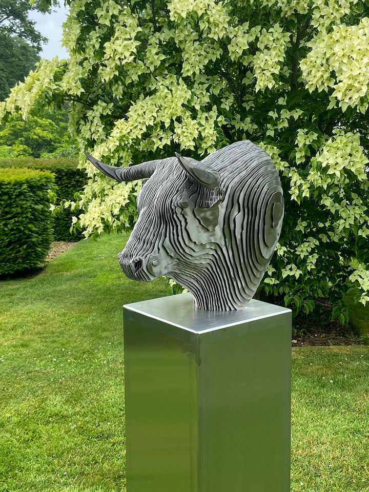 A bold stainless steel sculpture of a bull’s head by Tom Hiscox, composed of layered contour slices with curved horns and muscular form, set against vivid autumn foliage in an outdoor garden setting.