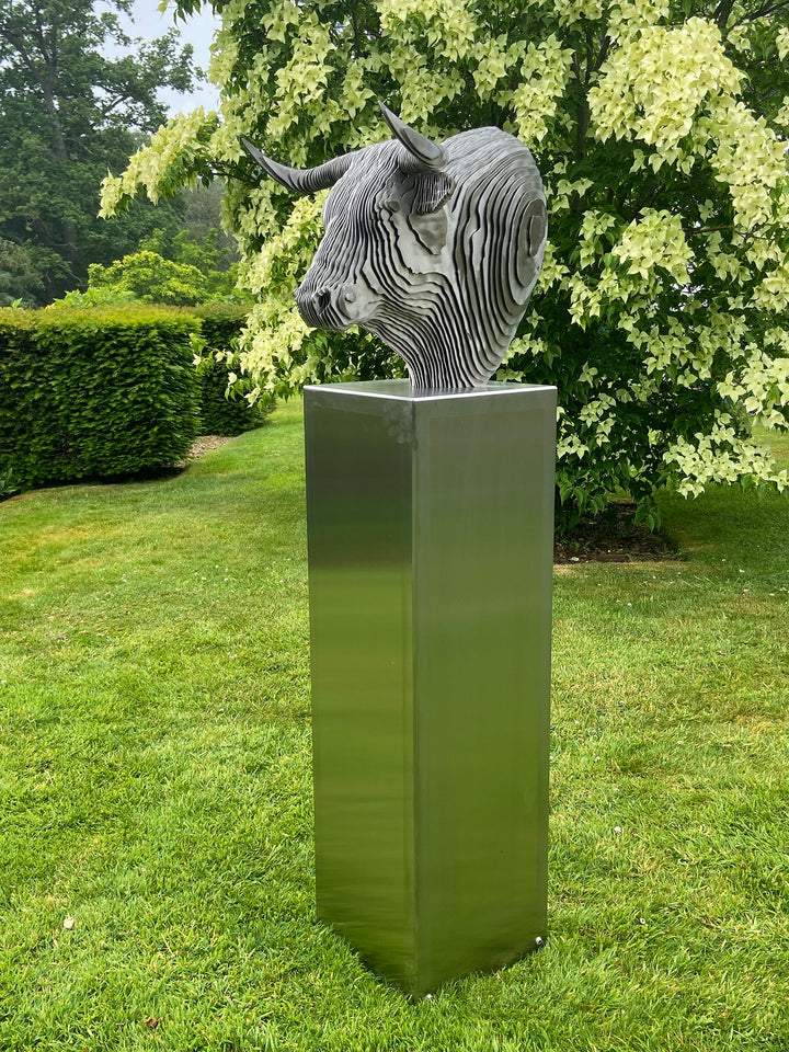 A bold stainless steel sculpture of a bull’s head by Tom Hiscox, composed of layered contour slices with curved horns and muscular form, set against vivid autumn foliage in an outdoor garden setting.