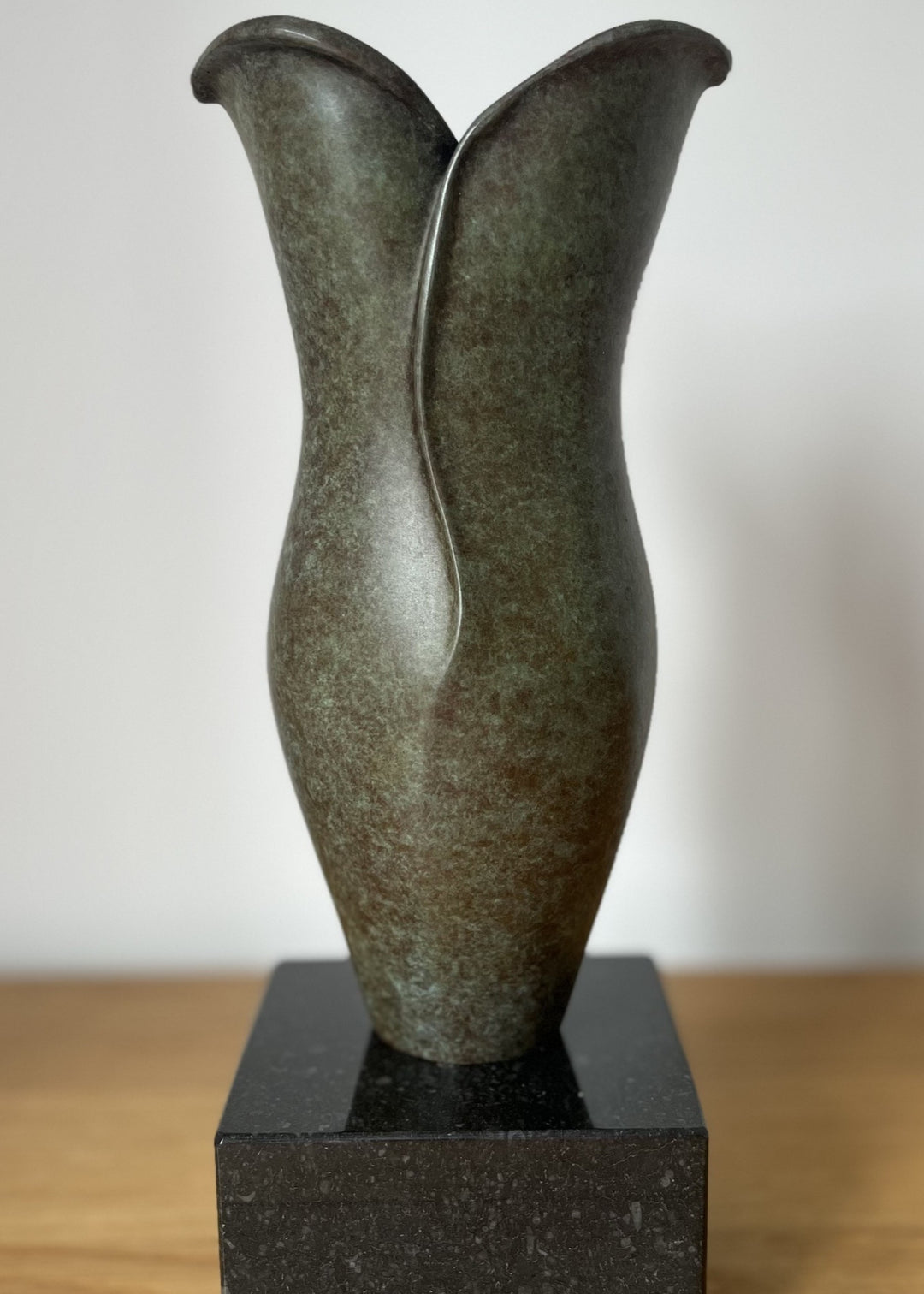 Small bronze sculpture with soft, organic curves that rise and part at the top like unfolding petals, finished in a mottled green and earthy brown patina, set on a polished black stone base.