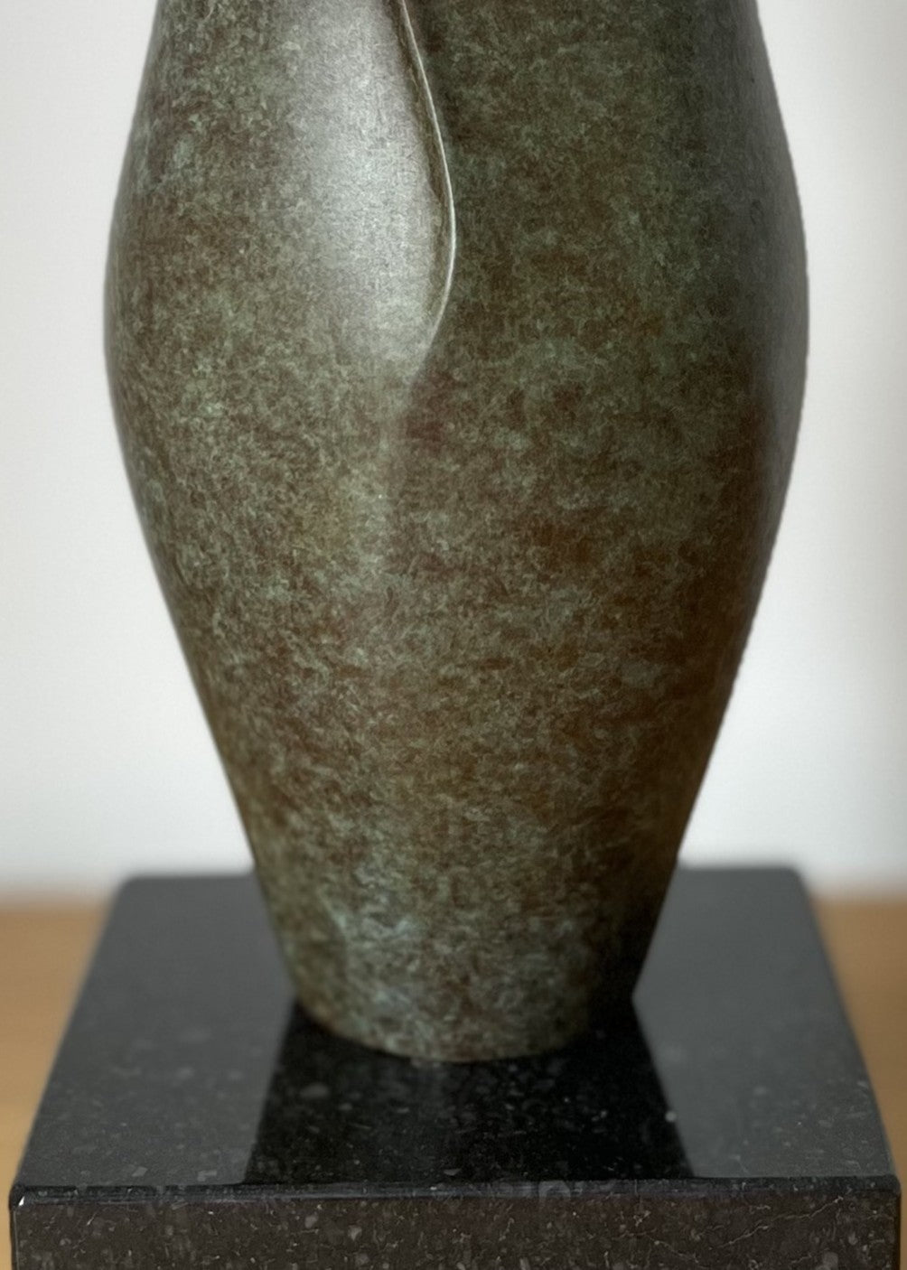 Small bronze sculpture with soft, organic curves that rise and part at the top like unfolding petals, finished in a mottled green and earthy brown patina, set on a polished black stone base.