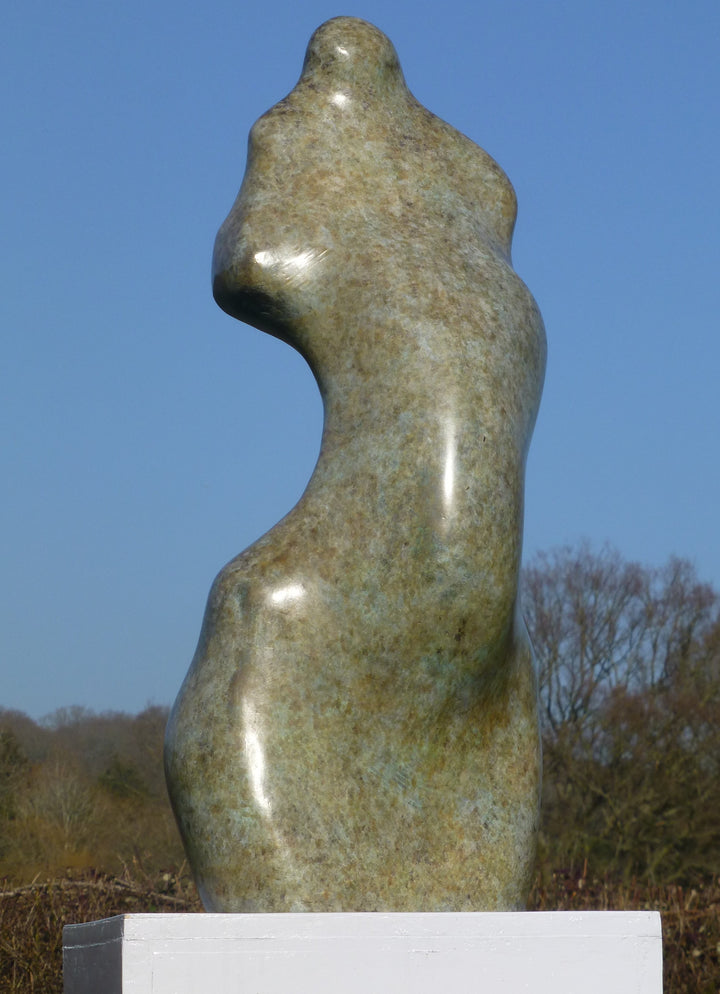 Bronze sculpture of an abstract female torso by Nicola Godden, with smooth organic curves and a softly polished surface, standing on a plinth outdoors against a clear blue sky, the form evoking the human body through simplified, flowing contours and natural balance.
