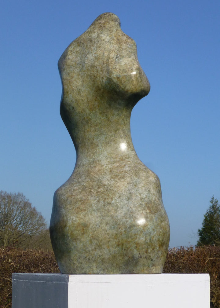Bronze sculpture of an abstract female torso by Nicola Godden, with smooth organic curves and a softly polished surface, standing on a plinth outdoors against a clear blue sky, the form evoking the human body through simplified, flowing contours and natural balance.
