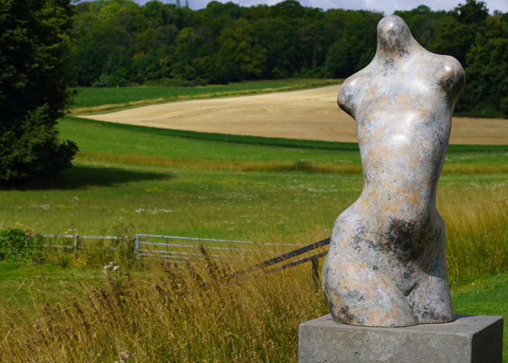Bronze abstract female torso sculpture by Nicola Godden, with softly curved organic form and smooth hand-finished surface, standing on a plinth outdoors, evoking sensuality, balance and metamorphosis through simplified human anatomy and tactile patination.