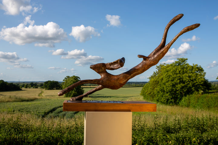 Freedom Majestic Hare sculpture by Min Reid, dynamic leaping hare captured mid-flight in warm bronze toned iron resin, mounted on a wooden base with countryside landscape, green fields and blue sky with white clouds in the background