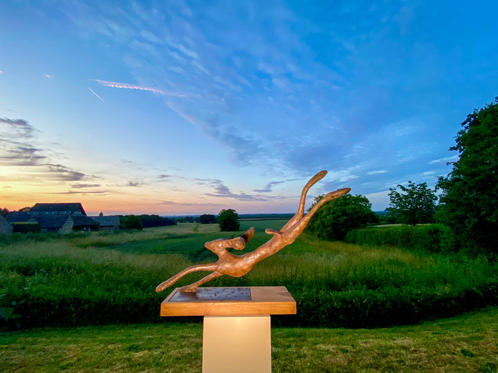 Freedom Majestic Hare sculpture by Min Reid, dynamic leaping hare captured mid-flight in warm bronze toned iron resin, mounted on a wooden base with countryside landscape, green fields and blue sky with white clouds in the background