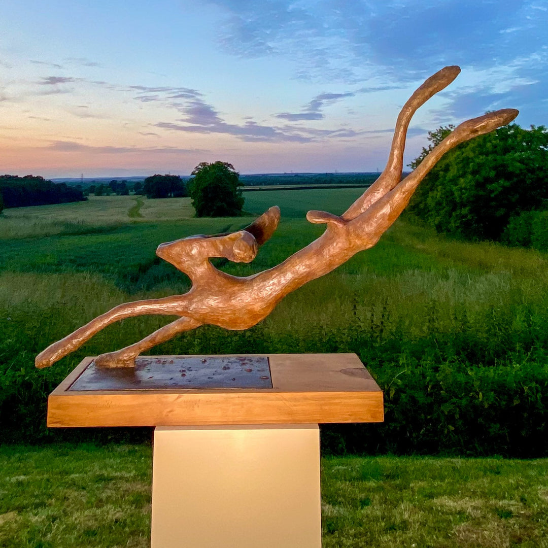 Freedom Majestic Hare sculpture by Min Reid, dynamic leaping hare captured mid-flight in warm bronze toned iron resin, mounted on a wooden base with countryside landscape, green fields and blue sky with white clouds in the background