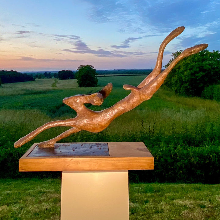 Freedom Majestic Hare sculpture by Min Reid, dynamic leaping hare captured mid-flight in warm bronze toned iron resin, mounted on a wooden base with countryside landscape, green fields and blue sky with white clouds in the background