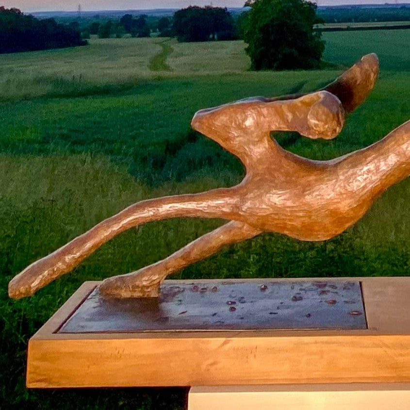Freedom Majestic Hare sculpture by Min Reid, dynamic leaping hare captured mid-flight in warm bronze toned iron resin, mounted on a wooden base with countryside landscape, green fields and blue sky with white clouds in the background