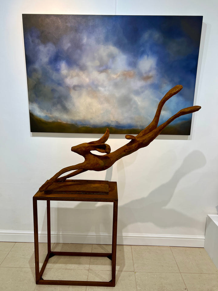 Freedom Majestic Hare sculpture by Min Reid, dynamic leaping hare captured mid-flight in warm bronze toned iron resin, mounted on a wooden base with countryside landscape, green fields and blue sky with white clouds in the background
